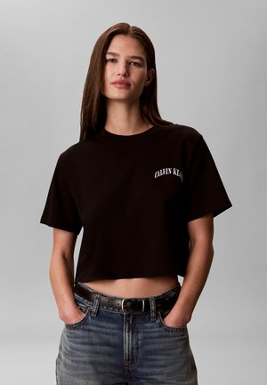 Femme aux longs cheveux bruns portant un t-shirt court noir Calvin Klein et un jean bleu, les mains dans les poches, fond gris.