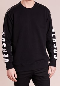 Sweat-shirt noir à manches longues, poignets côtelés et accents zippés sur les épaules. "VERSACE" imprimé en blanc le long de la manche gauche.