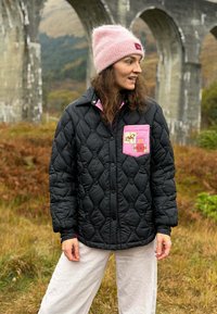 Žena oblečená v černé prošívané bundě s růžovou kapsou a růžovou pletenou čepicí stojí venku poblíž kamenného obloukového mostu na podzim.