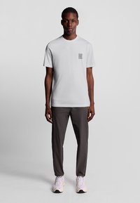 Lys grå t-shirt med et lille gråt logo på brystet, parret med mørkegrå bukser og lyse sneakers. Glat bomuldsstof.