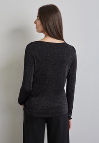 Maglietta nera a maniche lunghe con una texture scintillante, scollo rotondo e design attillato. Indossata con pantaloni neri, mostrando una silhouette fluida.