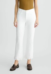 Pantalones vaqueros de mezclilla blanca de corte acampanado con un dobladillo con bordado de encaje. Combinados con una blusa marrón y náuticos oscuros.