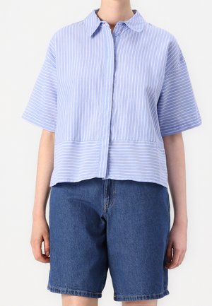Personne portant une chemise à manches courtes rayée bleu clair avec boutons et un short en denim bleu foncé, debout, bras détendus.