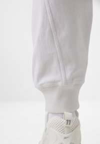 Világosszürke joggingnadrág, bordás mandzsettákkal és oldalsó varrással, fehér sneakerrel párosítva, amely hálós részletekkel és számmal ellátott sarokkal rendelkezik.
