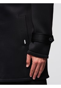 Main à côté de la manche d'un manteau noir avec un détail de sangle boutonnée, près d'une poche avec une petite étiquette métallique sur un fond propre.