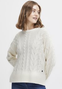 Pulover tricotat de culoare crem, cu un model de cablu, margini late cu nervuri și mâneci lungi. Prezintă un mic emblema neagră în apropierea colțului stâng de jos.