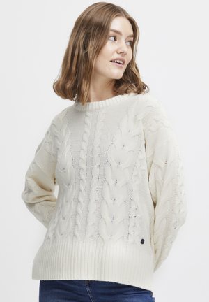 Cremfarbener, gestrickter Pullover mit Zopfmuster, breitem Rippbund und langen Ärmeln. Hat ein kleines schwarzes Emblem nahe der unteren linken Ecke.