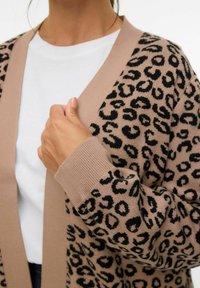 Cardigan en imprimé léopard beige et noir. Matière en tricot doux, design à ouverture frontale, poignets côtelés, texture subtile, associé à un t-shirt blanc.