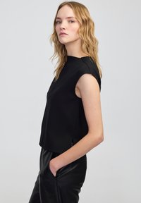 Schwarzes ärmelloses Top mit geradem Schnitt, das an den Seiten subtile Schlitze aufweist. Der Stoff sieht glatt und leicht strukturiert aus.