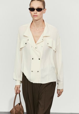 Femme portant une veste blanche avec des boutons argentés et un pantalon foncé, tenant un sac à main marron et portant des lunettes de soleil foncées, marchant vers l'avant.