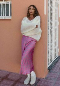 Woman leaning against peach wall wearing white oversized sweater, pink patterned skirt, white socks, and gray clogs on tiled sidewalk.