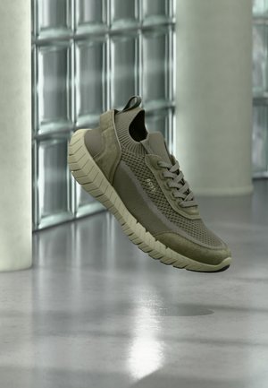 Baskets en maille texturée vert olive avec semelle épaisse beige, lacets à l'avant et languette, flottant à l'intérieur près d'un mur en blocs de verre et d'un sol poli.