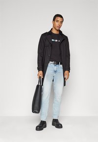 Abrigo negro sobre una camiseta negra, jeans de mezclilla azul claro, botas negras gruesas y un bolso tote negro. Diseño simple y colores sólidos.