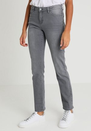 Personne portant un jean gris slim et des baskets blanches, debout devant un fond uni clair.