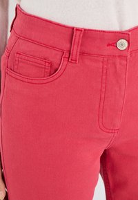 Vaqueros de mezclilla roja con una textura suave, diseño de cinco bolsillos y una forma ajustada. Cuenta con un cierre de botón metálico y detalle de costura en contraste.