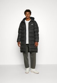 Crna pufasta jakna s kapuljačom, s prošivenim dizajnom, zatvaranjem na zip i logom na prsima. Uparena s sivim hlačama i bijelim tenisicama.