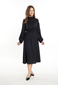 Rochie midi neagră cu mâneci lungi, talie strânsă și yoke texturat. Asortată cu pantofi negri, pe un fundal alb.