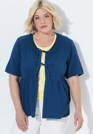 Kobieta o krótkich blond włosach, ubrana w niebieski krótki kardigan na żółtym topie i białych spodniach, stojąca na białym tle.