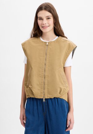 Beige mouwloze vest met een rits aan de voorkant, zijzakken en een ronde kraag. Gemaakt van zacht, lichtgewicht materiaal. Gedaagd over een wit shirt.