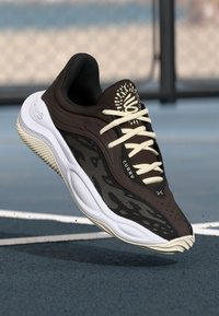 Sort og hvid Curry basketballsko med lys beige snørebånd og sål, i luften over en bane med sløret hegn i baggrunden.