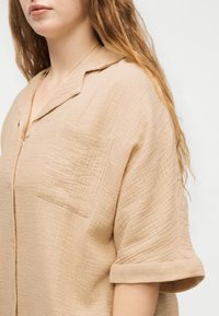 Γυναίκα με μπεζ υφασμάτινο πουκάμισο με κουμπιά, τσέπη στο στήθος και σηκωμένα κοντά μανίκια, με κυματιστά ανοιχτό καστανά μαλλιά ορατά.