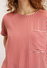 Kvinna iklädd röd- och vitrandig skjorta med liten ficka på vänster sida och uppvikt korta ärmar.