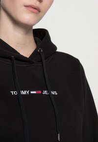 Nära bild på en person som bär en svart hoodie med "TOMMY JEANS" och en broderad röd, vit och blå flagglogotyp på bröstet.