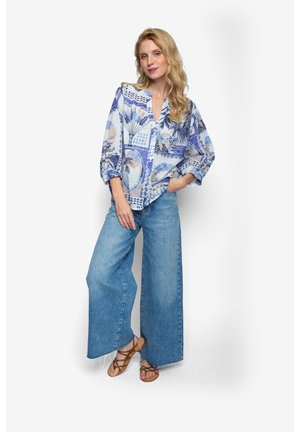 Fließende, hellblaue Bluse mit abstrakten tropischen Mustern und V-Ausschnitt, kombiniert mit weit geschnittenen, hellblauen Jeans und braunen Sandalen.