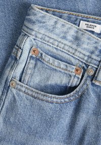 Jean en denim bleu clair avec une coupe décontractée, présentant des clous en cuivre, des coutures contrastantes et un petit détail de poche avant.