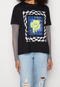 T-shirt nero con una grafica che presenta un volto sorridente giallo, circondato da testo blu e un bordo a scacchi. Indossato sopra una maglietta a maniche lunghe in rete.