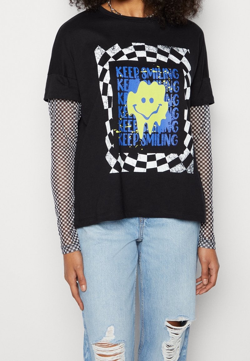 T-shirt nero con una grafica che presenta un volto sorridente giallo, circondato da testo blu e un bordo a scacchi. Indossato sopra una maglietta a maniche lunghe in rete.
