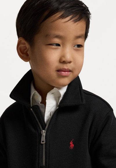 Schwarzer Reißverschluss-Pullover mit hohem Kragen, versehen mit einem roten Polo-Logo. Getragen über einem weißen Hemd mit Kragen, hergestellt aus strukturiertem Strickmaterial.