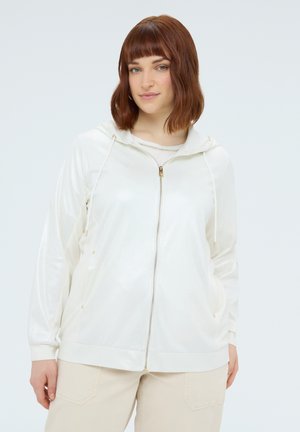 Junge Frau mit schulterlangem kastanienbraunem Haar, die einen weißen Kapuzenpullover mit Reißverschluss und beigefarbene Hosen trägt und vor einem hellen Hintergrund steht.
