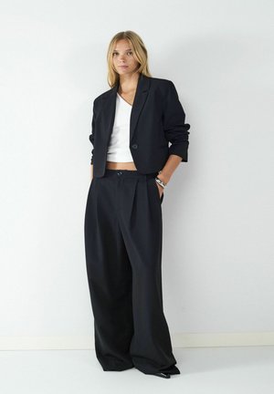 Blazer noir court avec un unique bouton et un large pantalon noir. Porté par-dessus un haut blanc à col en V, avec un tissu lisse et des lignes épurées.