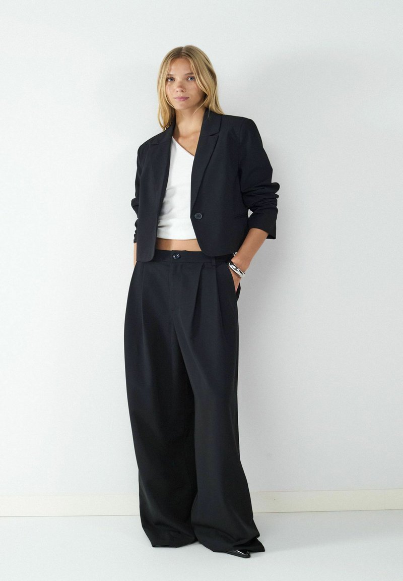 Czarna krótką marynarka z pojedynczym guzikiem i szerokimi czarnymi spodniami. Noszona na białej bluzce z dekoltem w kształcie litery V, wykonana z gładkiego materiału i o prostych liniach.