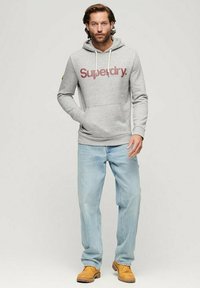 Felpa grigia in cotone con logo "Superdry" in bordeaux, tasche frontali, cappuccio regolabile con coulisse. Abbinata a jeans in denim chiaro e stivali gialli.