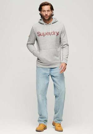 Grijze katoenen hoodie met het "Superdry" logo in bordeauxrood, voorzakken, capuchon met trekkoord. Gekoppeld aan lichtgewassen denim jeans en gele laarzen.
