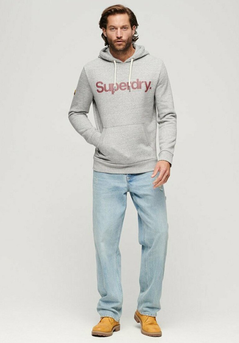 Felpa grigia in cotone con logo "Superdry" in bordeaux, tasche frontali, cappuccio regolabile con coulisse. Abbinata a jeans in denim chiaro e stivali gialli.