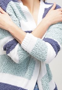 Cardigan tricoté à rayures horizontales en vert menthe, bleu marine et blanc. Tissu texturé avec une apparence chaleureuse et moelleuse, et un design ouvert à l'avant.