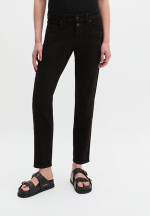 Person, die eng anliegende schwarze Jeans und schwarze Sandalen mit goldenen Schnallen trägt, vor einem schlichten hellen Hintergrund stehend.