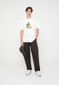 Maglietta bianca in cotone con un design grafico colorato, abbinata a pantaloni scuri e larghi e un cappello grigio, su uno sfondo neutro.