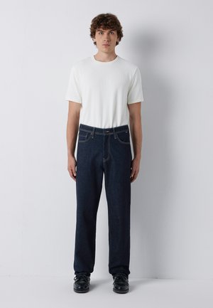 Young man standing against white wall wearing a white short-sleeve shirt, dark blue jeans, and black lace-up shoes.