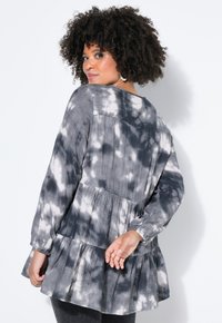 Grijs-wit tie-dye top met een gelaagde peplumstijl, lange mouwen en een ronde halslijn. De stof lijkt zacht met subtiele textuurdetails.