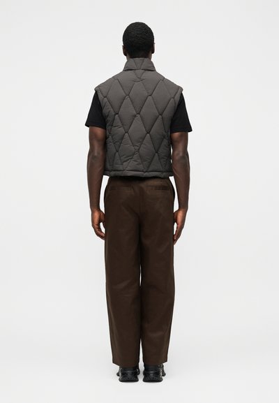 Colete cinzento acolchoado com gola alta sobre uma t-shirt preta, combinado com calças castanhas de perna larga e sapatos pretos, visto por trás.