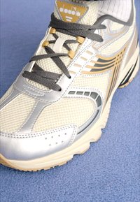 Fehér és ezüst atletikai cipő, hálós felsőrésszel, szürke fűzőszerkezettel és arany díszítésekkel; párnázott talppal rendelkezik és jellegzetes dizájnelemek jellemzik.