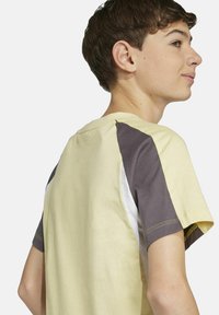 Figurnah geschnittenes T-Shirt mit kurzen Ärmeln in Hellgelb, mit grauen und weißen Schulterpaneelen. Aus Baumwollmaterial mit sichtbaren Nähakzenten.