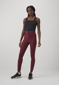 Čierny crop top s hranatým výstrihom a detailom s nastaviteľnou šnúrkou v kombinácii s vysokými bordovými legínami; model má na sebe čierne športové topánky.