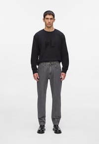 Sudadera negra con logo texturizado en el frente, combinada con jeans grises. El modelo lleva zapatos negros tipo slip-on. Diseño simple y moderno.