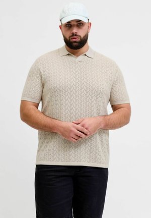 Uomo con polo beige a maniche corte con motivo e cappellino bianco, in piedi con le mani giunte, su sfondo chiaro uniforme.