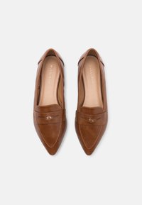 Mocassins en cuir marron avec un bout pointu, présentant une sangle décorative avec des éléments en or et des détails de couture. Texture lisse, design minimaliste.